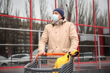 Tıbbi maskeli ve süpermarketin yanında alışveriş arabası olan olgun bir adam. Koronavirüs salgını kavramı
