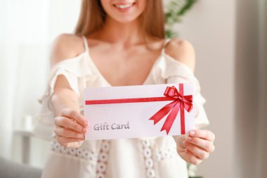 Beautiful young woman with gift card at home clipart