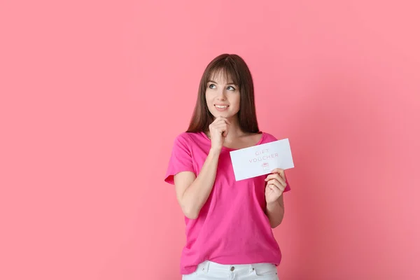 Thoughtful young woman with gift voucher on color background