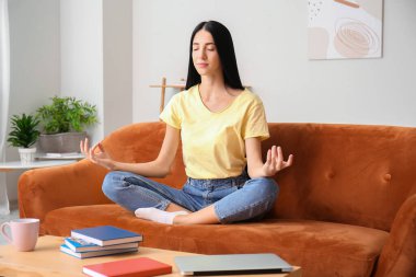Evdeki kanepede meditasyon yapan genç bir kadın.