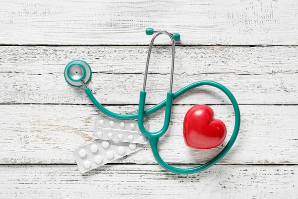 Modern stethoscope, pills and heart on light wooden background