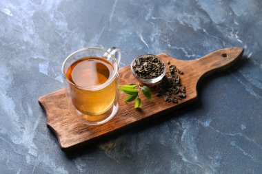 Board with cup of green tea and dry leaves on color background