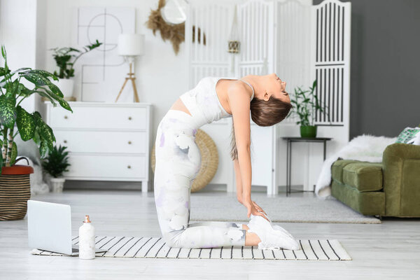 Sporty young woman with laptop practicing yoga at home during lockdown