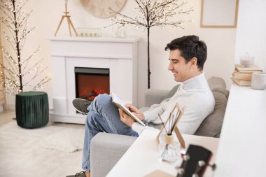 Evde şöminenin yanında kitap okuyan yakışıklı bir adam.