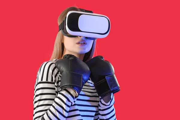 Young woman in boxing gloves and with virtual reality glasses on color background