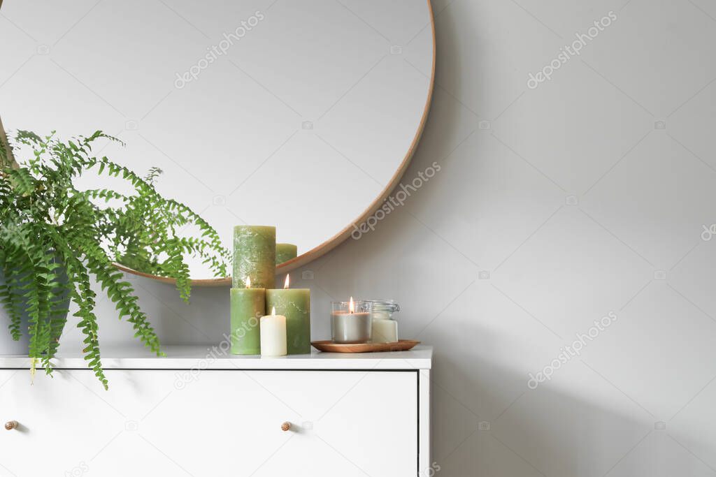 Burning candles and houseplant on chest of drawers in room