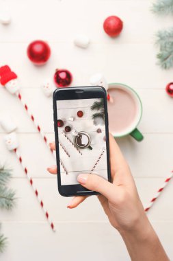 Woman taking photo of cup with hot cacao drink and Christmas decor on white wooden background
