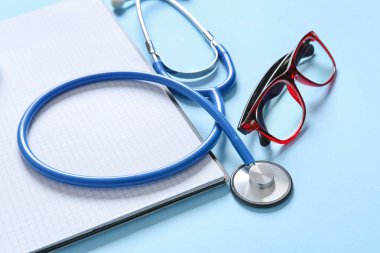 Modern stethoscope, notebook and eyeglasses on color background, closeup