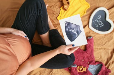 Yatakta sonogram görüntüsü ve bebek kıyafetleri olan hamile bir kadın.