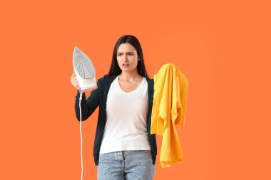 Upset young woman with iron and clothes on color background
