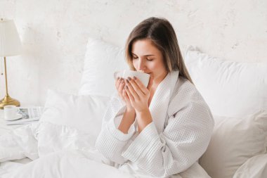 Genç bir kadın yatak odasında kahve içiyor.