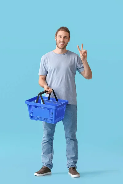 Junger Mann Mit Leerem Einkaufskorb Zeigt Siegesgeste Auf Farbigem Hintergrund — Stockfoto