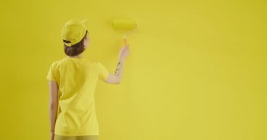 Sarı duvarın yanında boya silindirli genç bir kadın.