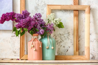 Işık duvarının yanındaki rafta leylak çiçekli vazolar var.