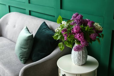 Odanın duvarının yanındaki masada leylak çiçekli vazo.