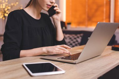 Evde dizüstü bilgisayar kullanan genç bir kadın cep telefonuyla konuşuyor.