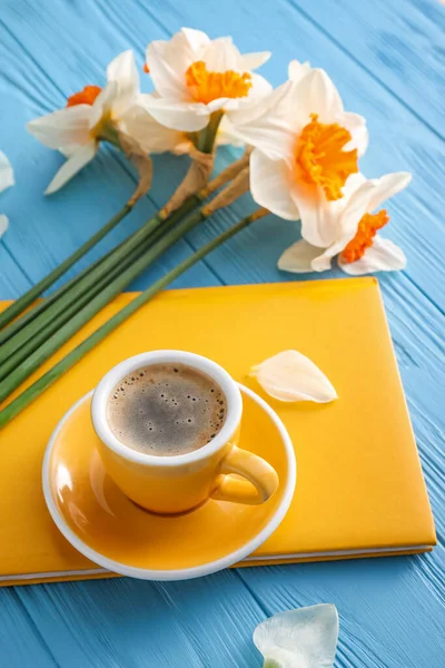 Cup of coffee, notebook and beautiful daffodils on color wooden background