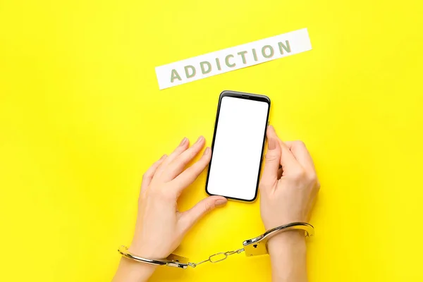 Woman with mobile phone and in handcuffs on color background. Concept of addiction to gadget