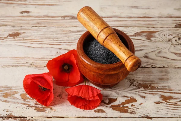 Mortar and pestle with poppy seeds and flowers on light wooden background