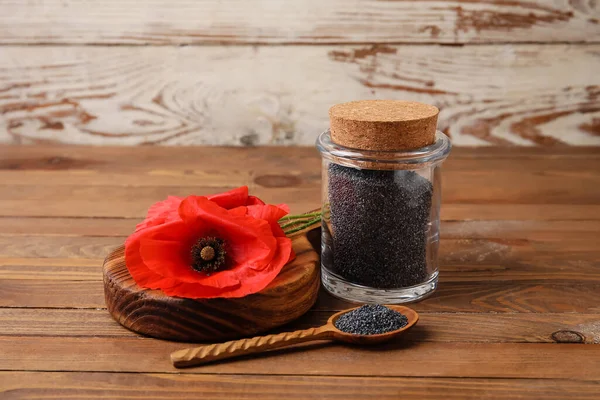 Jar and spoon with poppy seeds and flower on wooden background