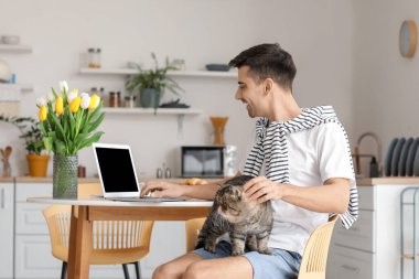 Mutfakta dizüstü bilgisayar kullanan sevimli kedisi olan genç bir adam.