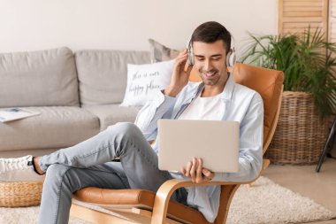 Kulaklıklı genç adam evde dizüstü bilgisayar kullanıyor.