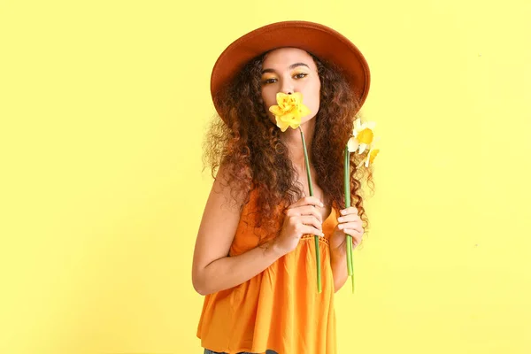 Beautiful young African-American woman with narcissi on color background