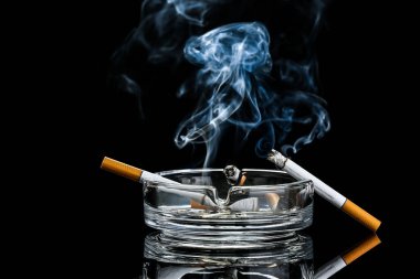 Ash tray with burning cigarettes on dark background clipart