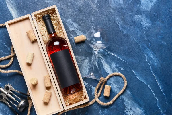 Box with bottle of wine, corkscrew and glass on color background