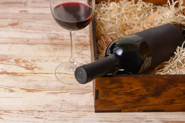Box with bottle of wine and glass on light wooden background, closeup