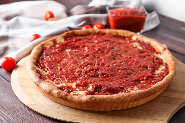 Board with tasty Chicago-style pizza, sauce and tomatoes on wooden background