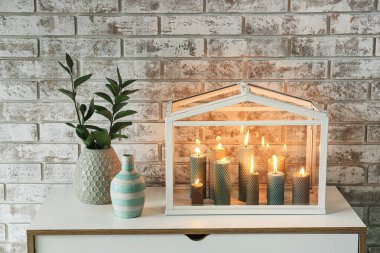 Tuğla duvarın yanındaki çekmecelerde yeşil dalları olan mumlar ve vazolar yakıyor.