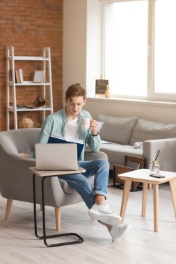 Genç adam kitap okuyor ve evdeki koltukta otururken kahve içiyor.