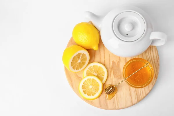 Glass bowl with sweet honey, lemon slices and teapot on wooden board against white background