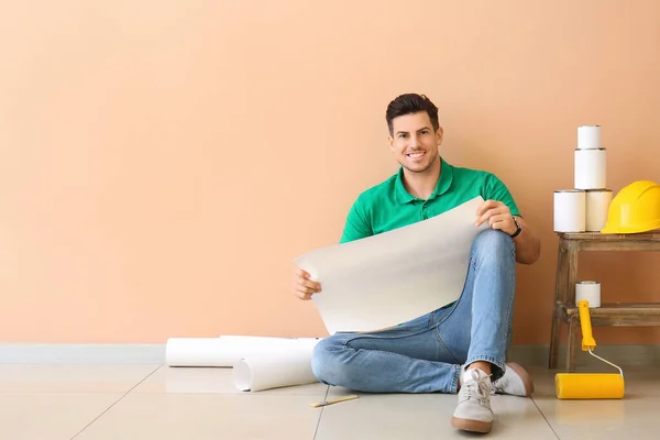 Young man with roller, house plan and cans of paint near color wall