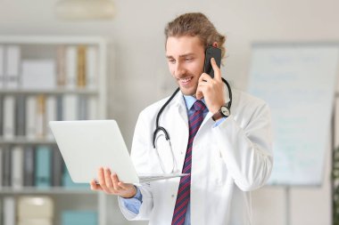 Bilgisayarlı genç doktor klinikte cep telefonuyla konuşuyor.