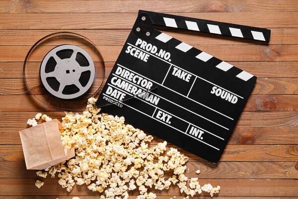 Tasty popcorn with movie clapperboard and reel on wooden background