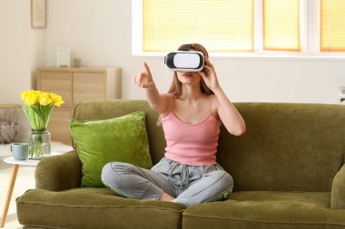 Evde sanal gerçeklik gözlüklü genç bir kadın var.