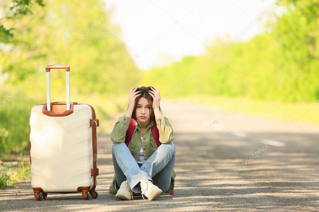 Upset young woman with suitcase hitchhiking on road