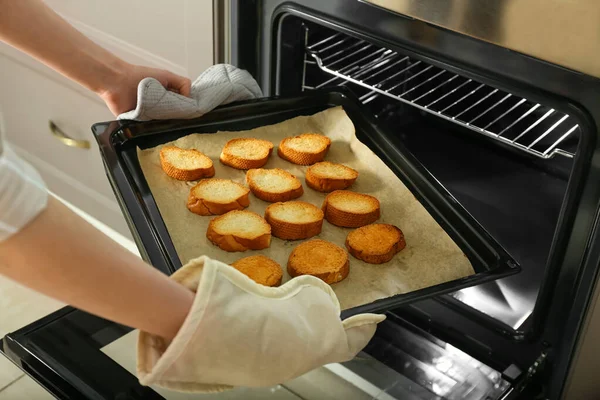 Woman taking baking sheet with toasted bread out of oven, closeup