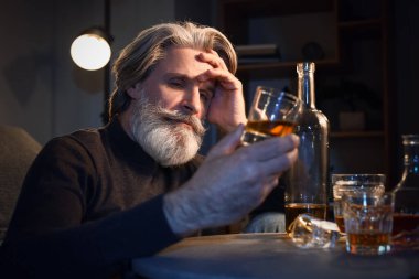 Drunk senior man with glass of whiskey late in evening at home