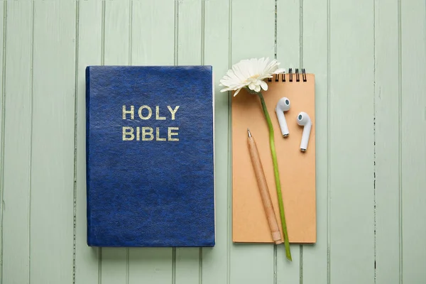 Holy Bible, earphones and flower on color wooden background