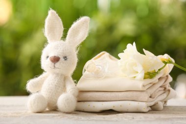 Dışarıdaki tahta masada bebek kıyafetleri, emzik, oyuncak ve çiçek yığını.