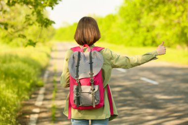 Sırt çantalı genç bir kadın yolda otostop çekiyor.