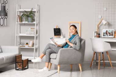 Elinde kahve ve dergi olan genç bir kadın evdeki koltukta oturuyor.