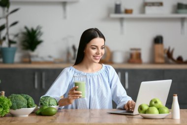 Young woman with glass of smoothie using laptop in kitchen