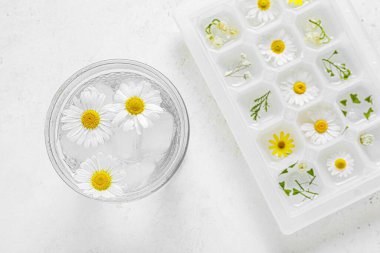 Bir bardak soğuk su ve buz tepsisi. Arka planda güzel papatya çiçekleri, yakın plan.