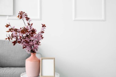 Vase with blossoming branches and blank frame on table near light wall