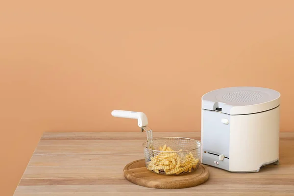 Modern deep fryer and tasty french fries on table near color wall