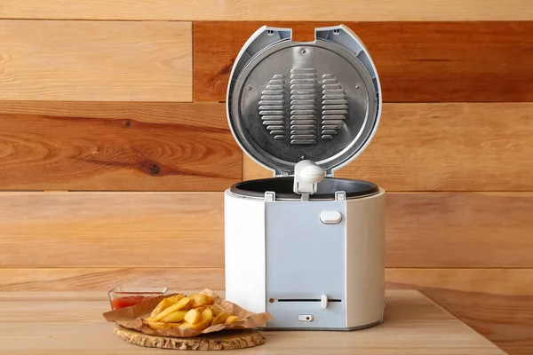 Modern deep fryer and tasty potato on table near wooden wall